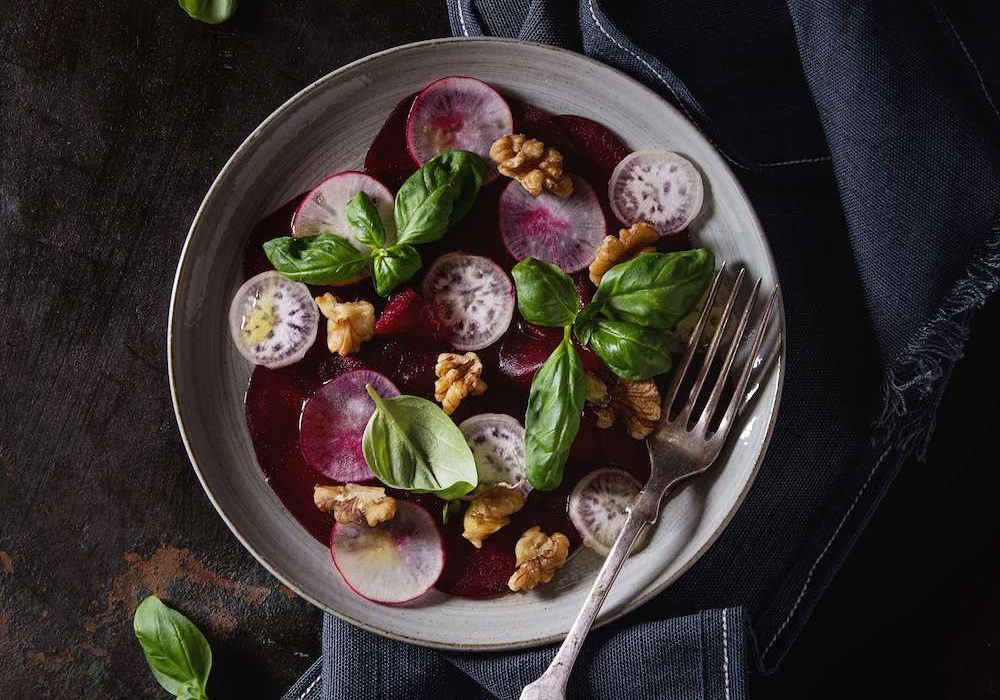 Vegan beetroot carpaccio salad with radish, basil, olive oil and walnuts in ceramic plate over dark rusty metal background. Top view, space. Dark rustic style, healthy eating