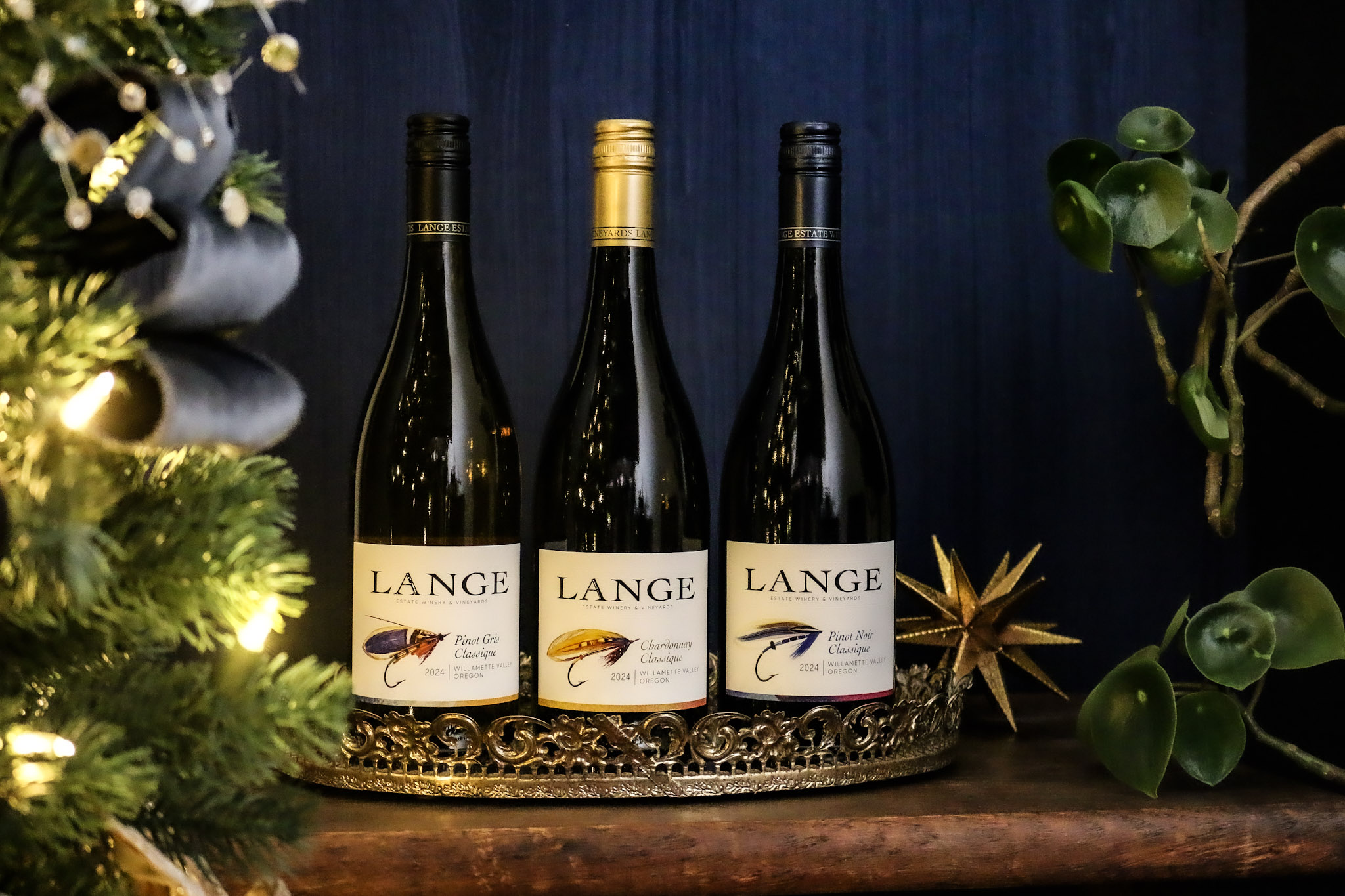 Three bottles, one each of Pinot Noir, Pinot Gris, and Chardonnay from Classique trio in front of blue background and Christmas tree