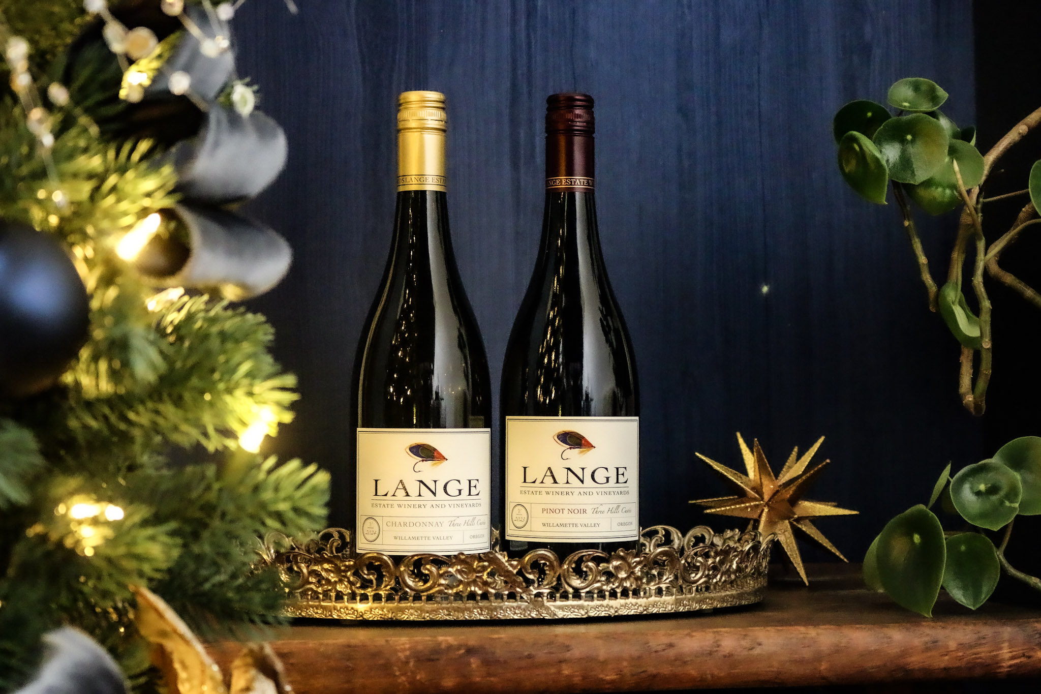 Two bottles of Three Hills Cuvee, one of chardonnay and one of Pinot Noir, in front of a blue background beside a Christmas tree
