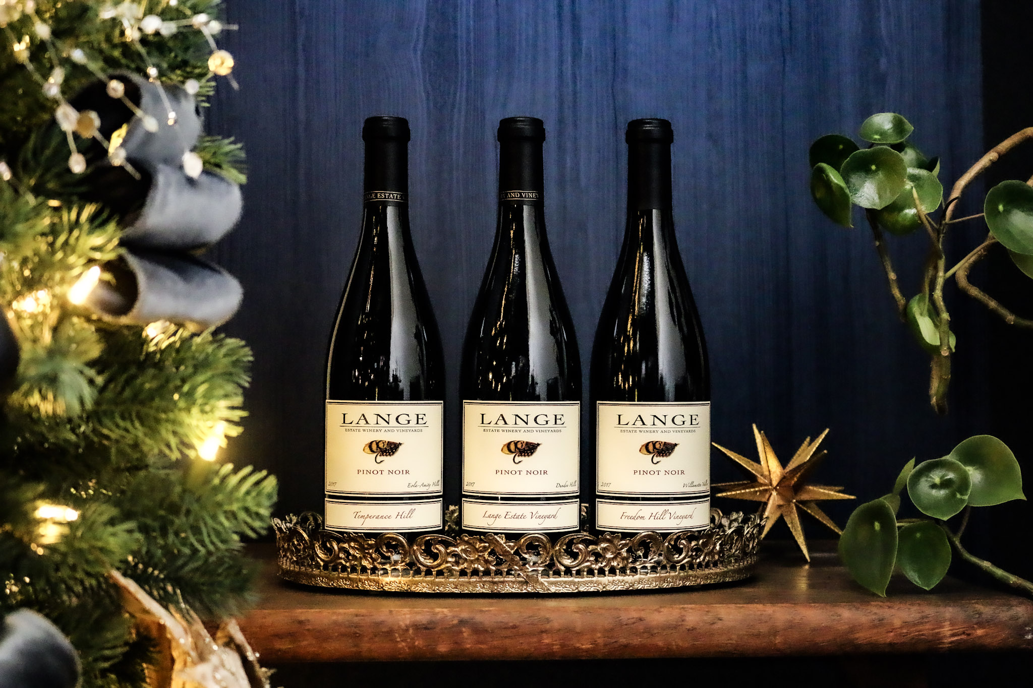 Three bottles of 2017 vintage Pinot Noir from Lange Estate, Freedom Hill, and Temperance Hill Vineyards in front of a blue background next to a Christmas tree
