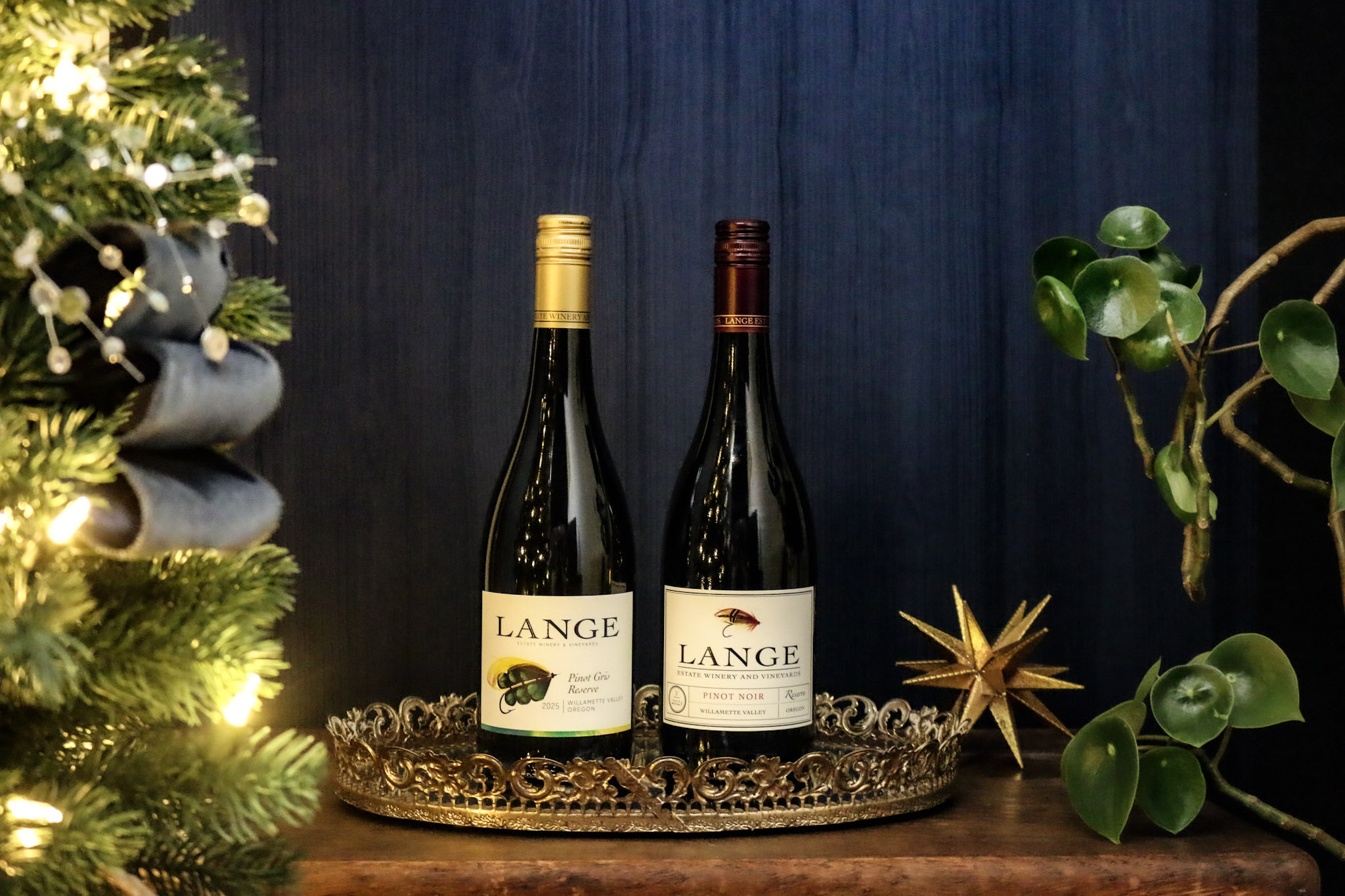 Two Bottles, one each of Reserve Pinot Noir and Reserve Pinot Gris, in front of a blue background next to a Christmas tree