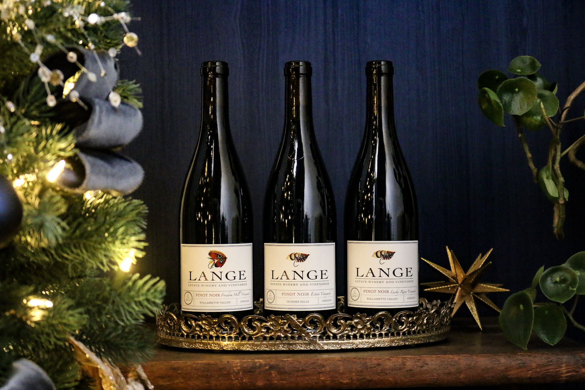 Three bottles of single-vineyard Pinot Noir, Lange Estate, Freedom Hill, and Lucky River Vineyard in front of a blue background next to a Christmas tree