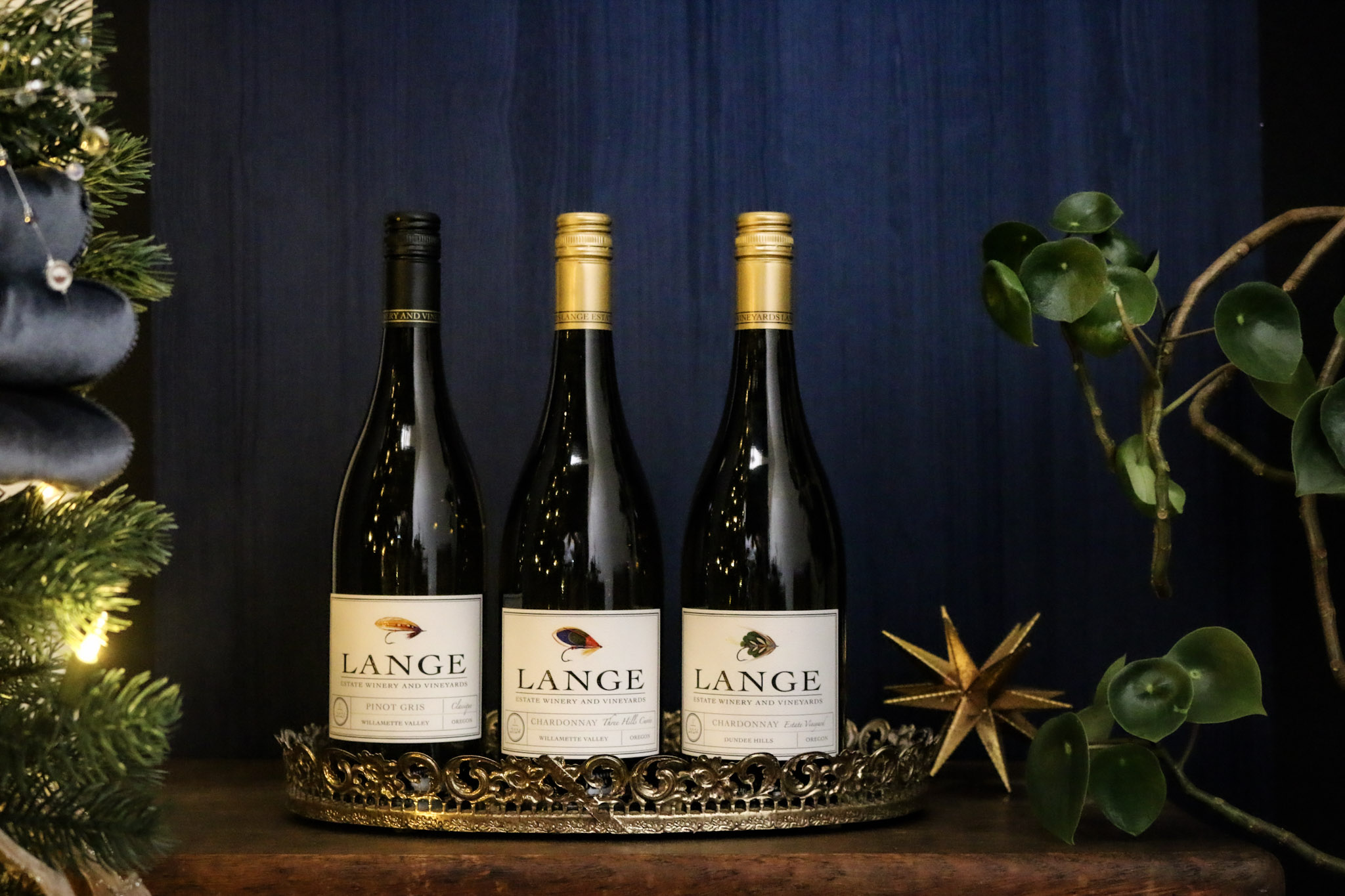 Three bottles of wine, Pinot Gris Classique, Estate Chardonay, and Three Hills Cuvee Chardonnay in front of a blue background next to a Christmas tree