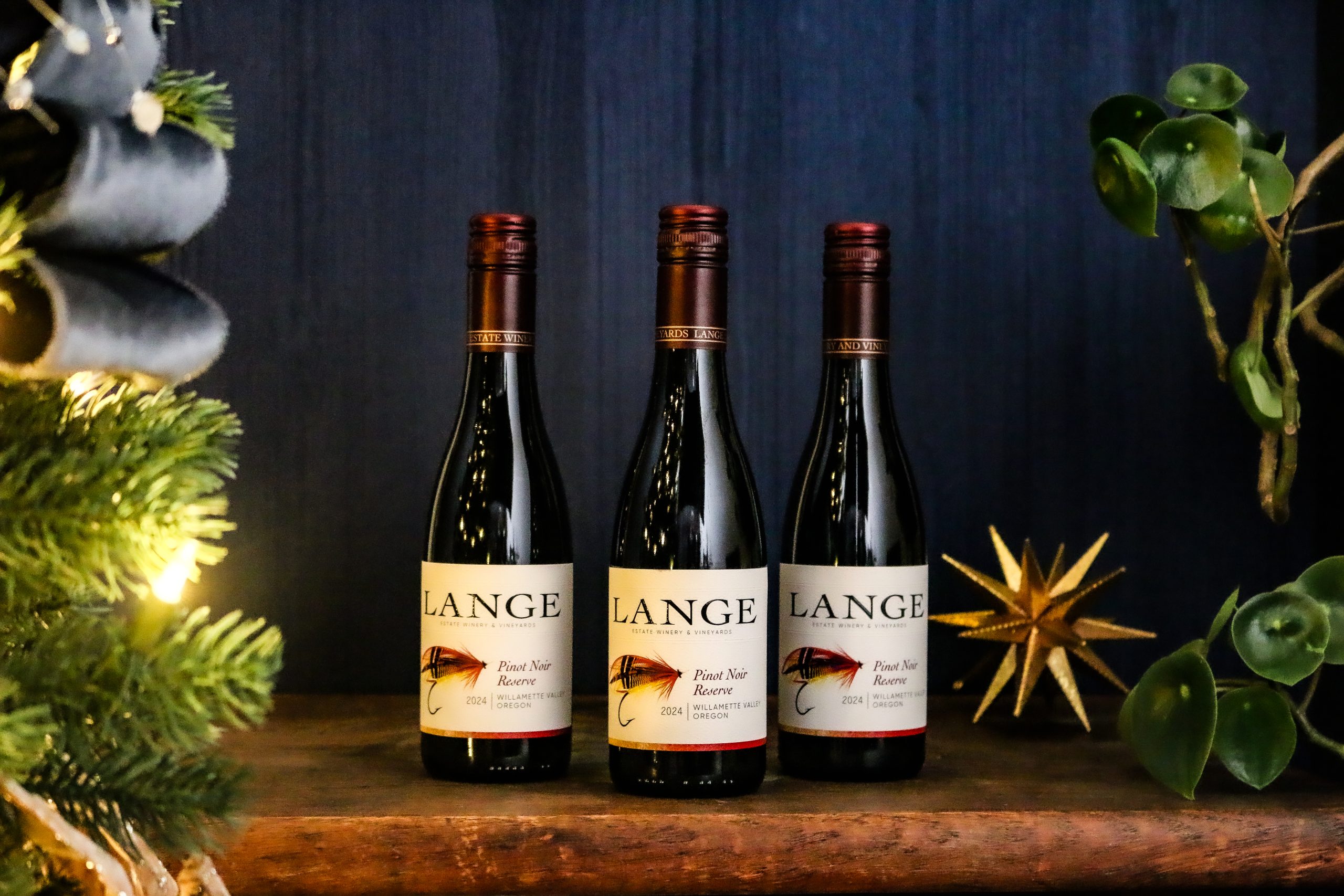 Three small 375ml bottles of Lange Pinot Noir Reserve in front of a blue background next to a Christmas tree and gold start